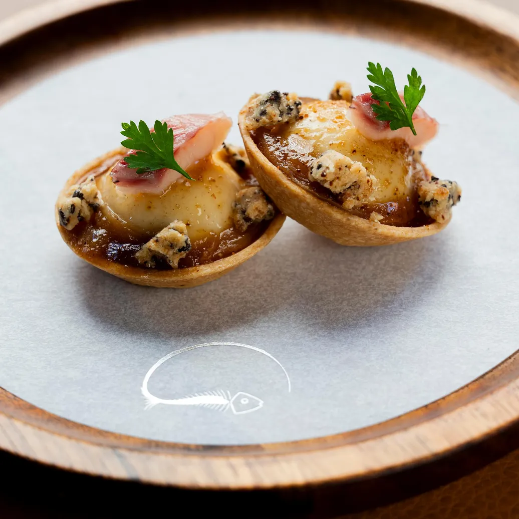 Deux petits pâtés garnis de crème et de morceaux de viande, décorés de fines tranches de radis et de feuilles de persil, présentés sur une assiette grise à bord en bois