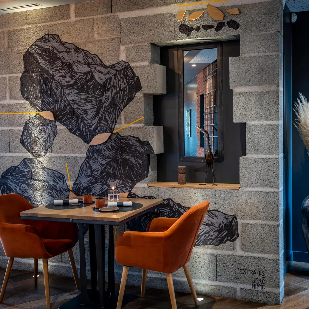 Table avec deux chaises orange dans un intérieur décoré de grandes feuilles murales et d'un vase avec des herbes sèches