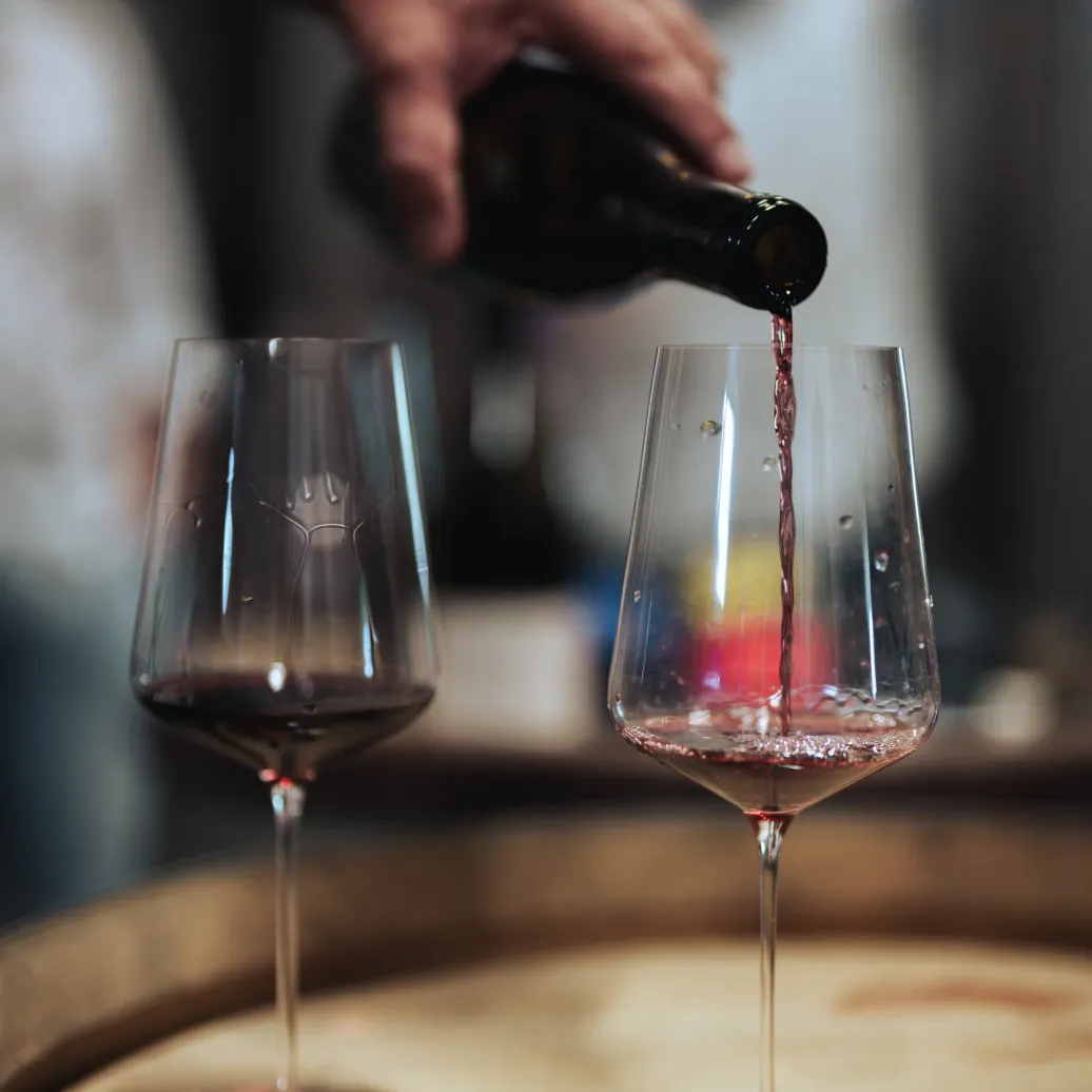 Une main verse du vin rouge dans un verre à pied posé sur une table en bois