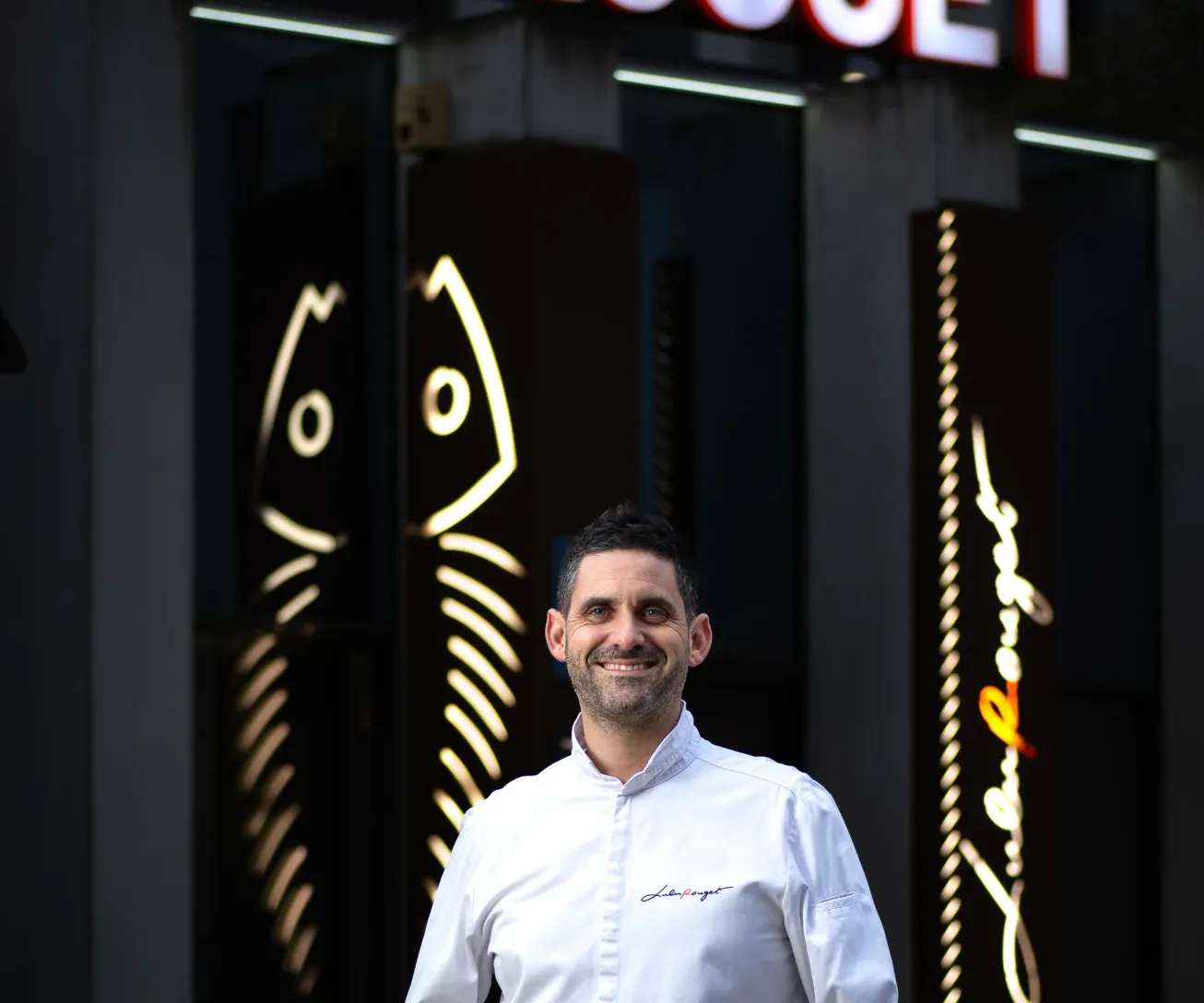 Un homme souriant en chemise blanche et jean devant l'enseigne lumineuse Lulu Rouge