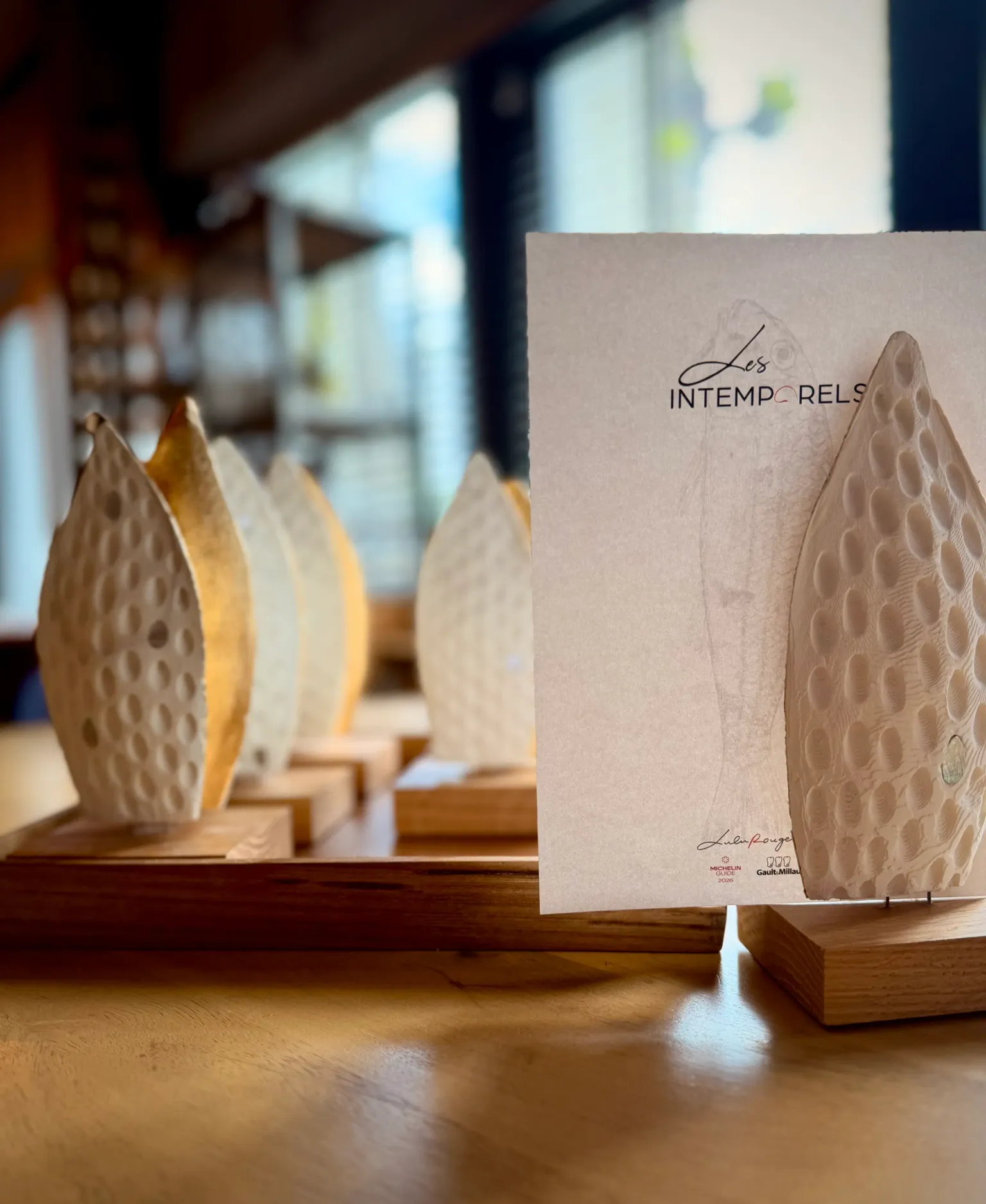 Des portes-menus en forme de feuilles sculptées sont disposés sur une table en bois avec une carte indiquant "Les Intemporels"